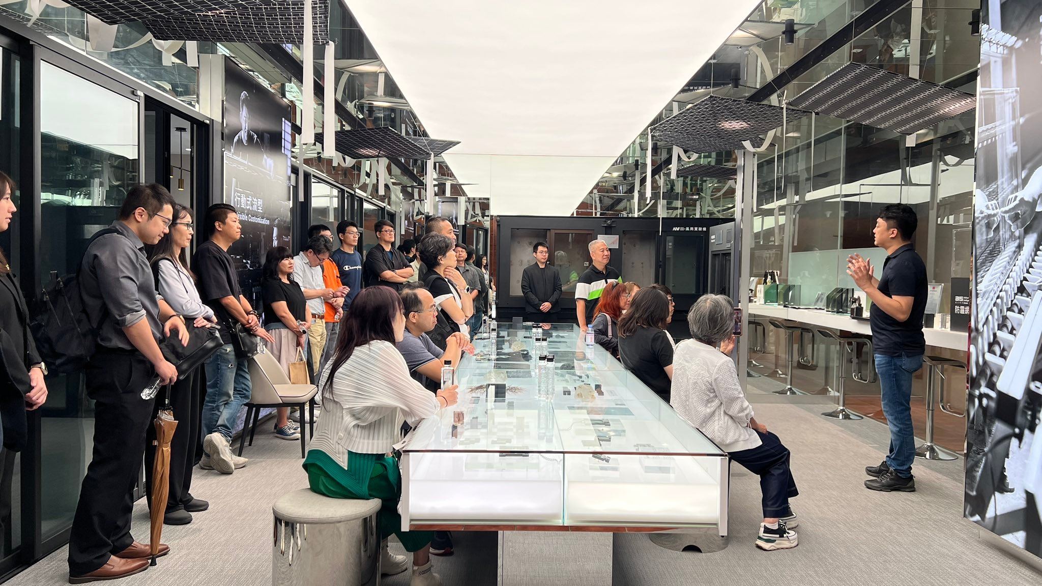 逢甲建設碩專系友會參訪老虎城旗艦店，成員圍在鋁型材與五金樣品展示櫃旁聽取導覽，背景為風雨實驗室測試艙